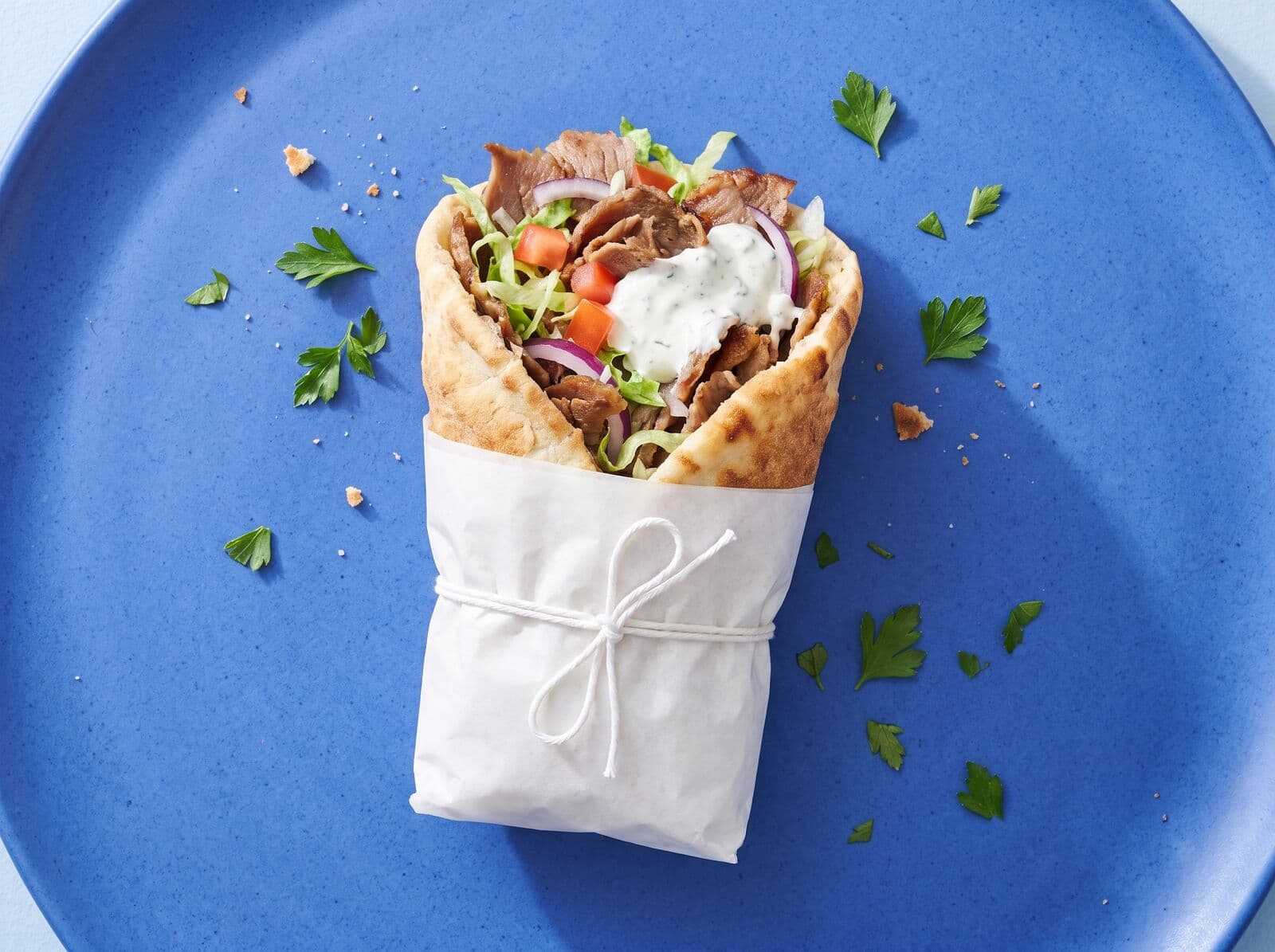 Beispielbild: Döner im Fladenbrot mit Salat, Tomate, Zwiebeln und heller Soße, in weißes Papier gewickelt, von oben auf blauem Teller